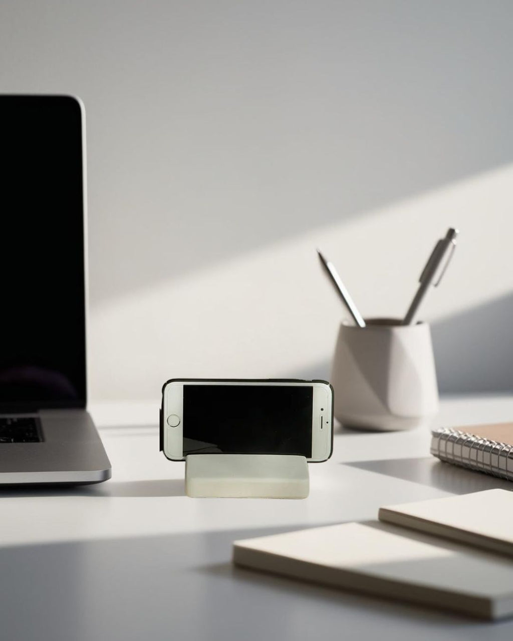White minimalist smartphone stand on modern desk with laptop, pen holder, and natural light