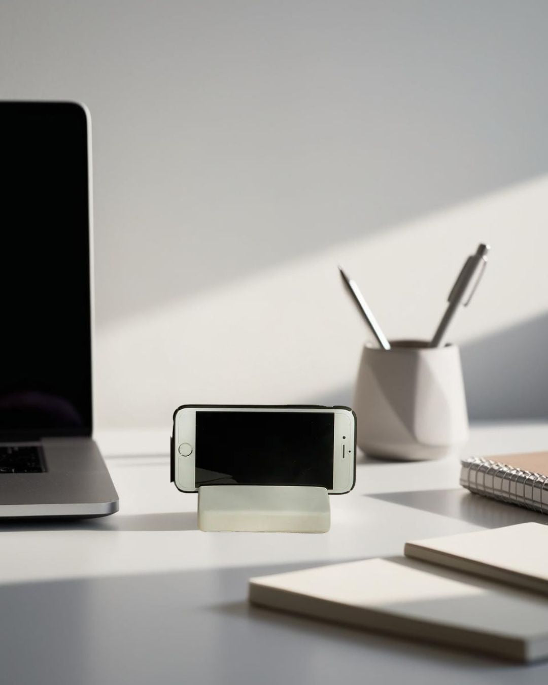 White minimalist smartphone stand on modern desk with laptop, pen holder, and natural light