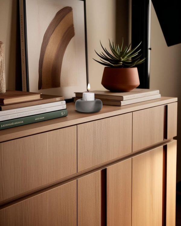 A white round concrete candle holder on a entry table, with a candle inside it, surrounded by books and a plant.
