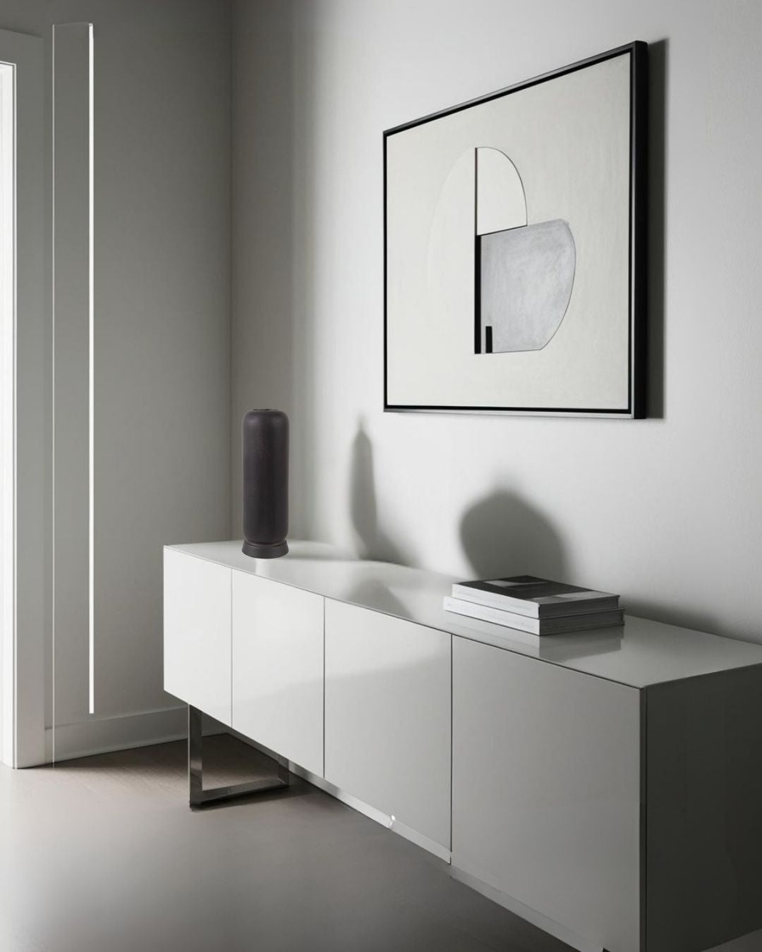 Modern wabi-sabi black cylindrical vase styled on a white glossy sideboard with minimalist abstract wall art and stacked books in a contemporary interior design.