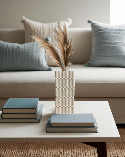 White geometric 3D printed vase with dried pampas grass on modern white coffee table in minimalist living room with beige sofa and blue cushions.






